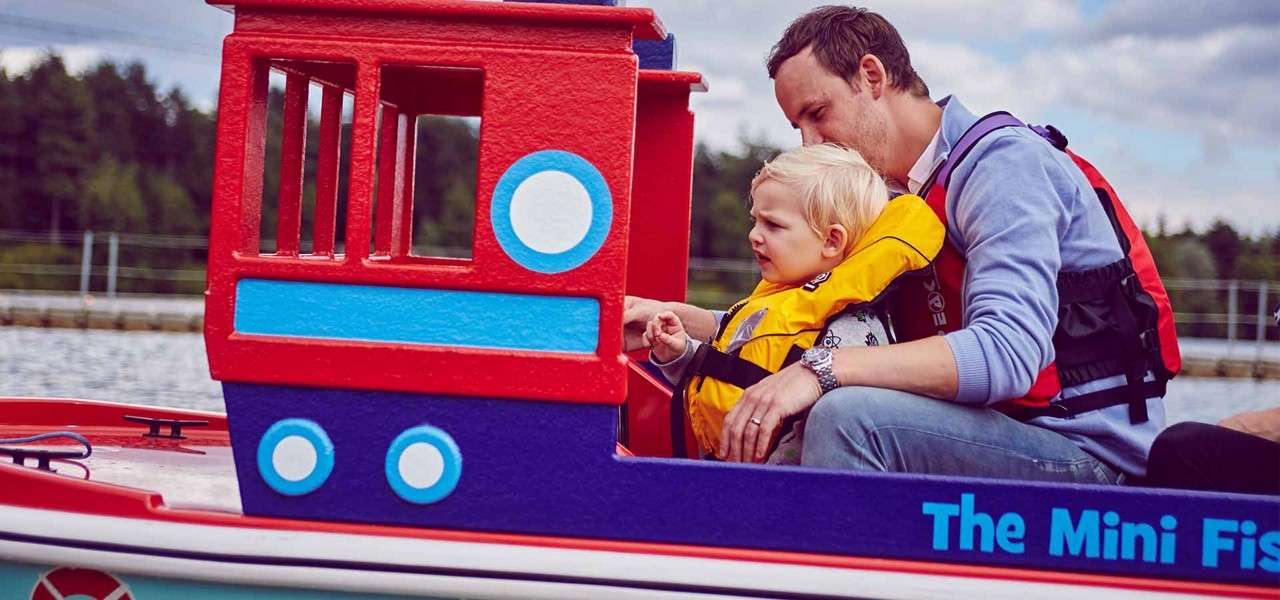 Child wearing a yellow life jacket steers a colorful mini boat while an adult supports them, on calm water near trees and docks.
Text: The Mini Fis