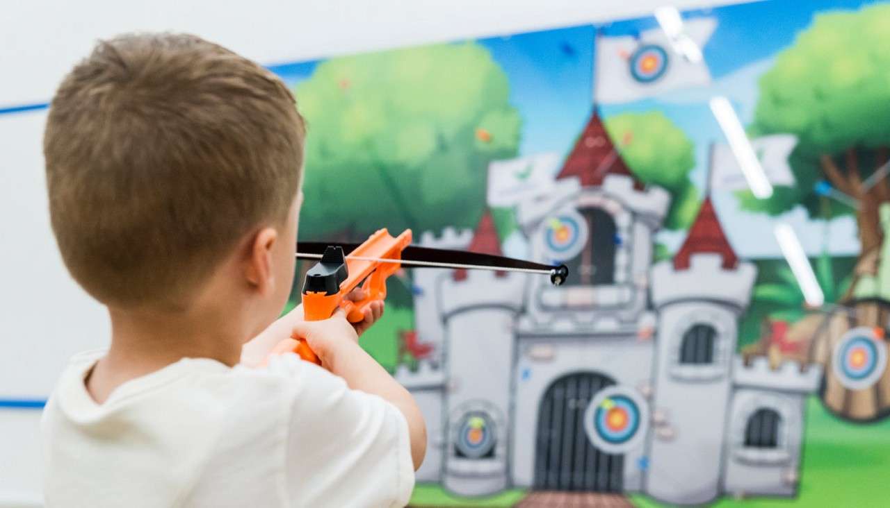Child holds an orange toy crossbow, aiming toward colorful bullseye targets on a cartoon castle backdrop, indoors with bright lighting in a playful, arcade-style environment.