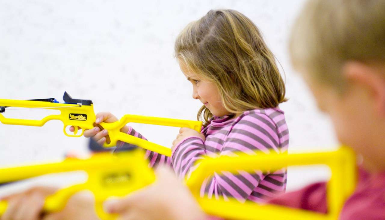 Children aim bright yellow toy rifles indoors, focusing down sights; one child in a striped shirt concentrates side-on, another blurred in the foreground, against a plain white wall background.