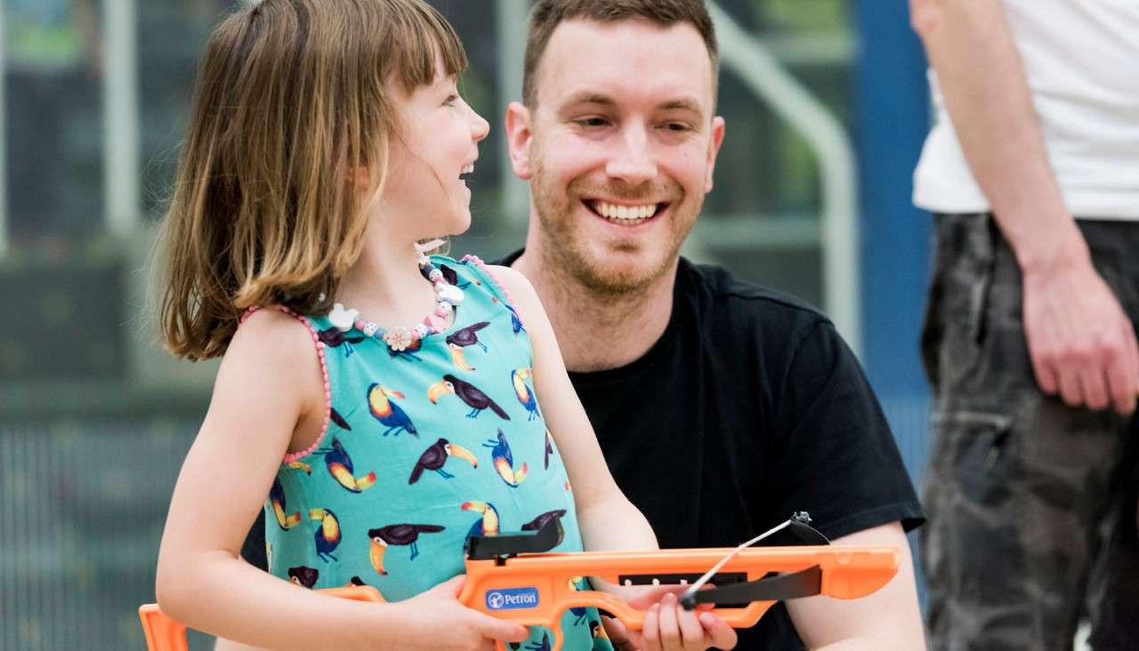 Child with orange toy crossbow holds and smiles toward a seated adult; indoors at a gym or recreation center, with another person standing nearby. Visible text: “Petron”.
