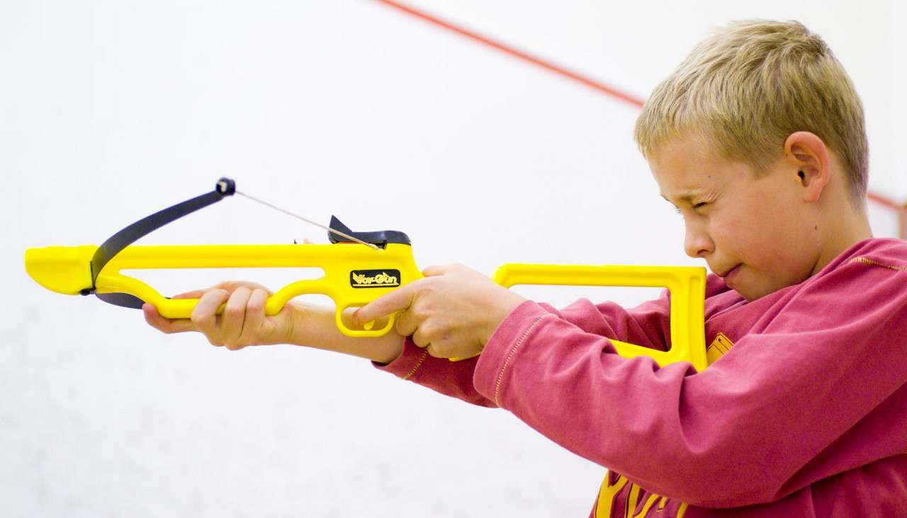 Boy aims a bright yellow toy crossbow, squinting as he pulls the trigger; indoors against a white wall with red lines. Text on the toy’s logo: “BowGun”.