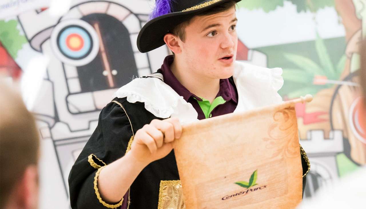 A young person in medieval costume holds a parchment scroll while speaking, against a cartoon castle and archery backdrop. Text on the scroll: "Center Parcs".