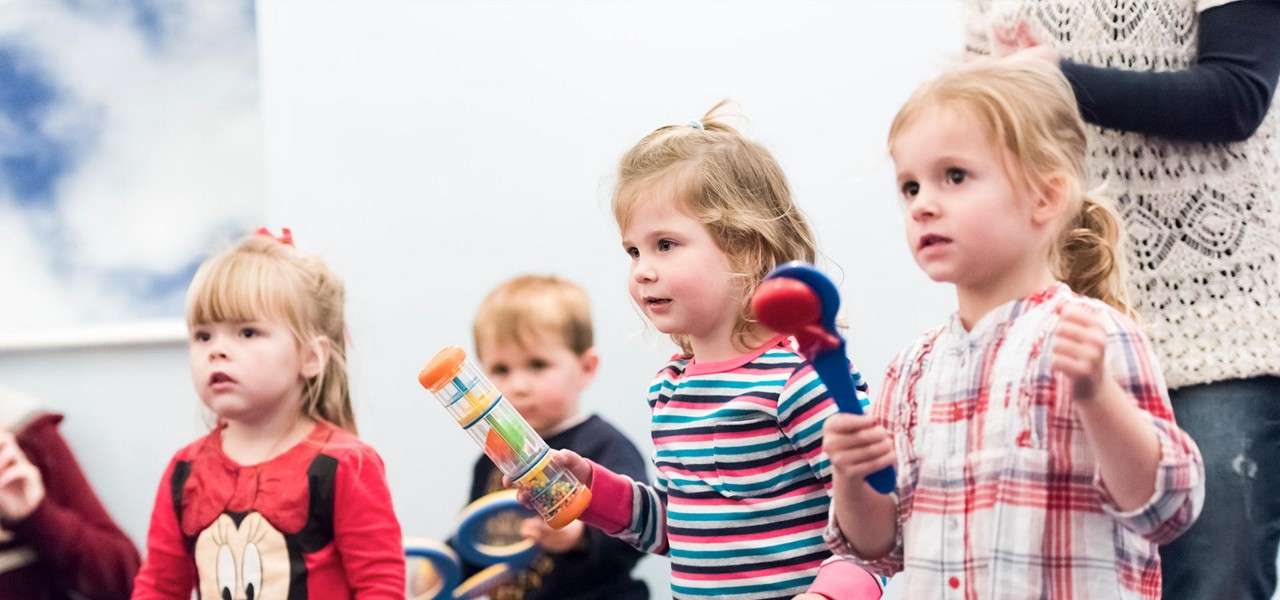 Three preschool children hold toy percussion instruments, shaking maracas and a rainstick, while standing in a bright classroom; two more children and an adult are in the background observing.