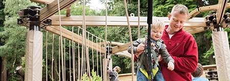 Child holds a rope while being supported by an adult, moving along a wooden rope-bridge structure in a playground; surrounding trees and beams frame the area.