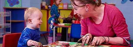 Toddler and caregiver finger-paint together, smearing colors on paper; both smile with paint-covered hands. In a bright daycare classroom, small tables, shelves, and another child painting in background.