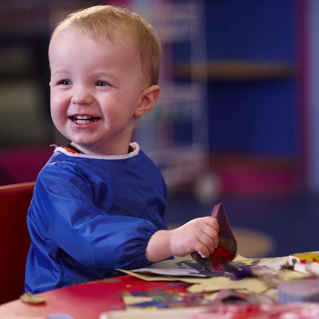 Toddler smiles while dabbing paint onto paper with a sponge, wearing a blue smock, seated at a craft table in a colorful classroom.