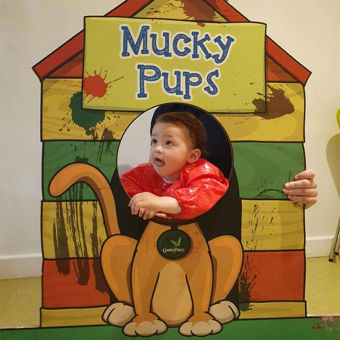 Child peeks through a round cutout, leaning forward in a red smock, inside a cartoon dog-house board. Surroundings show paint splatters and a hand steadying it. Text: “Mucky Pups”, “CenterParcs”.