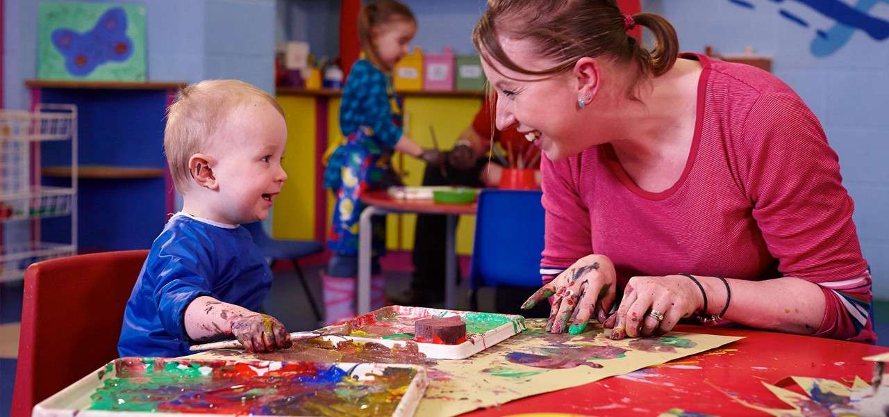 Child and caregiver finger-painting at a preschool table, smiling and chatting; hands smeared with bright colors; other children paint in the background amid colorful shelves and artwork.