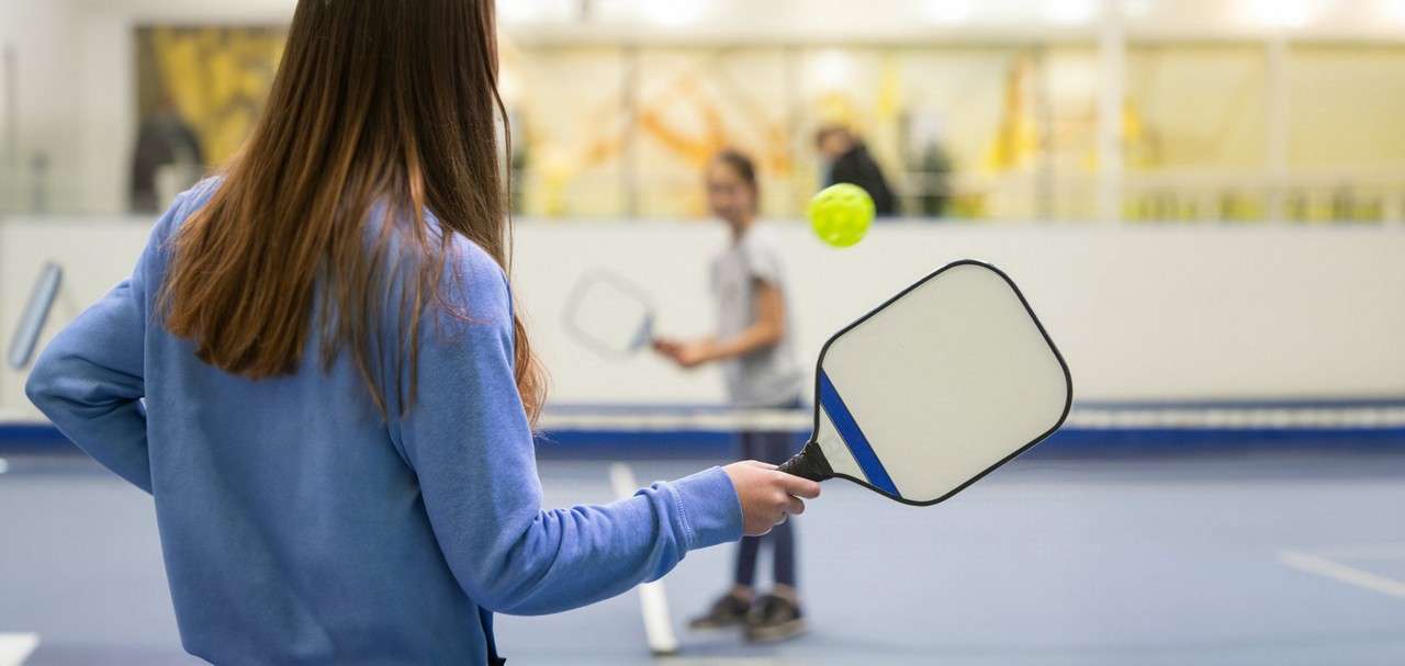 A person holding a paddle hits a yellow ball while playing pickleball on an indoor court, with another player across the net in the background.