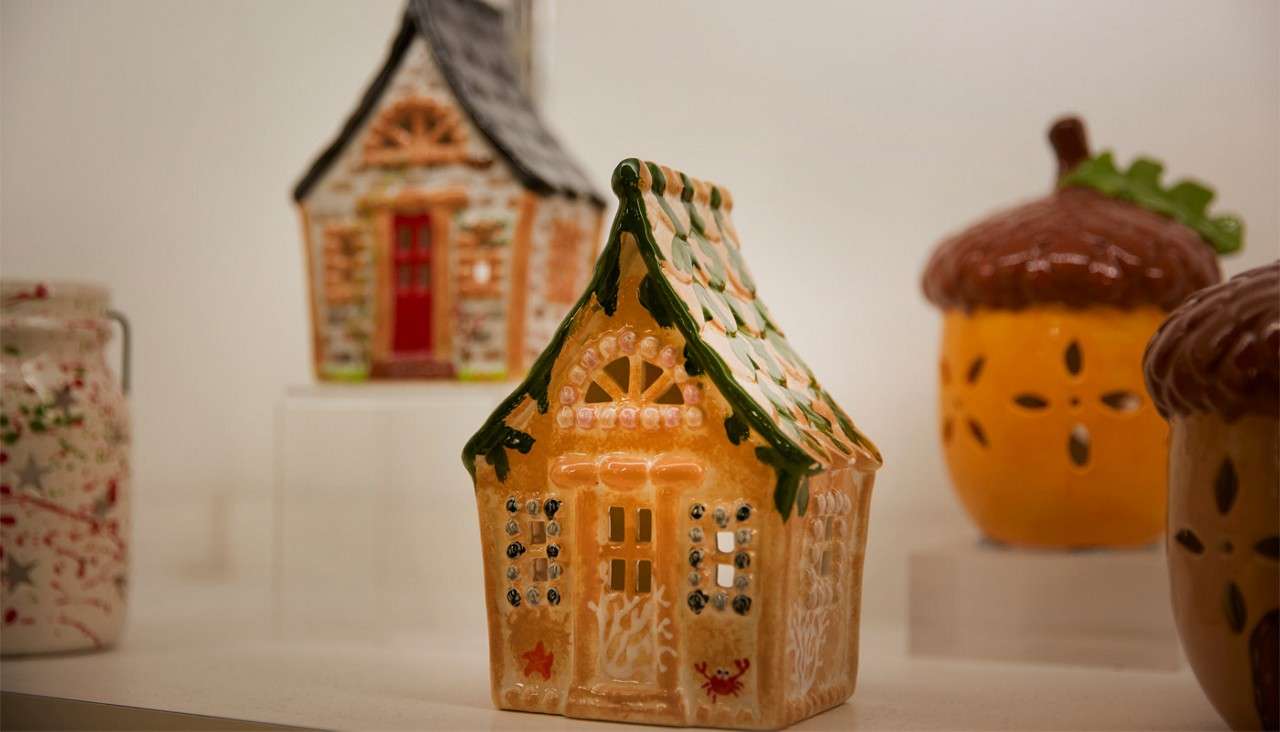 Ceramic house figurine sits on a display shelf; cutout windows and green-topped roof. Behind it, another house, acorn-shaped containers, and a speckled jar appear softly blurred.