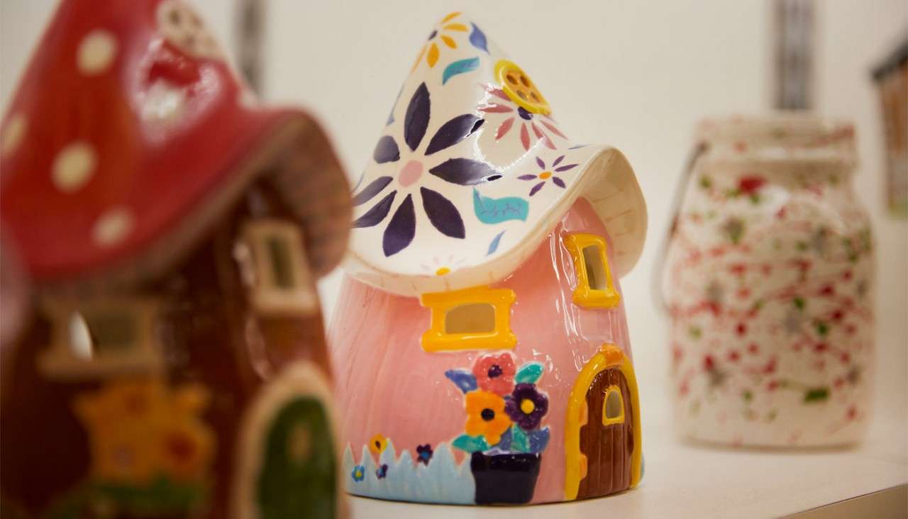 Ceramic fairy-house figurine stands upright, showing pink walls, yellow windows, a brown door, and a white flower-painted roof; context: displayed on a shelf among other blurred, decorative ceramics.