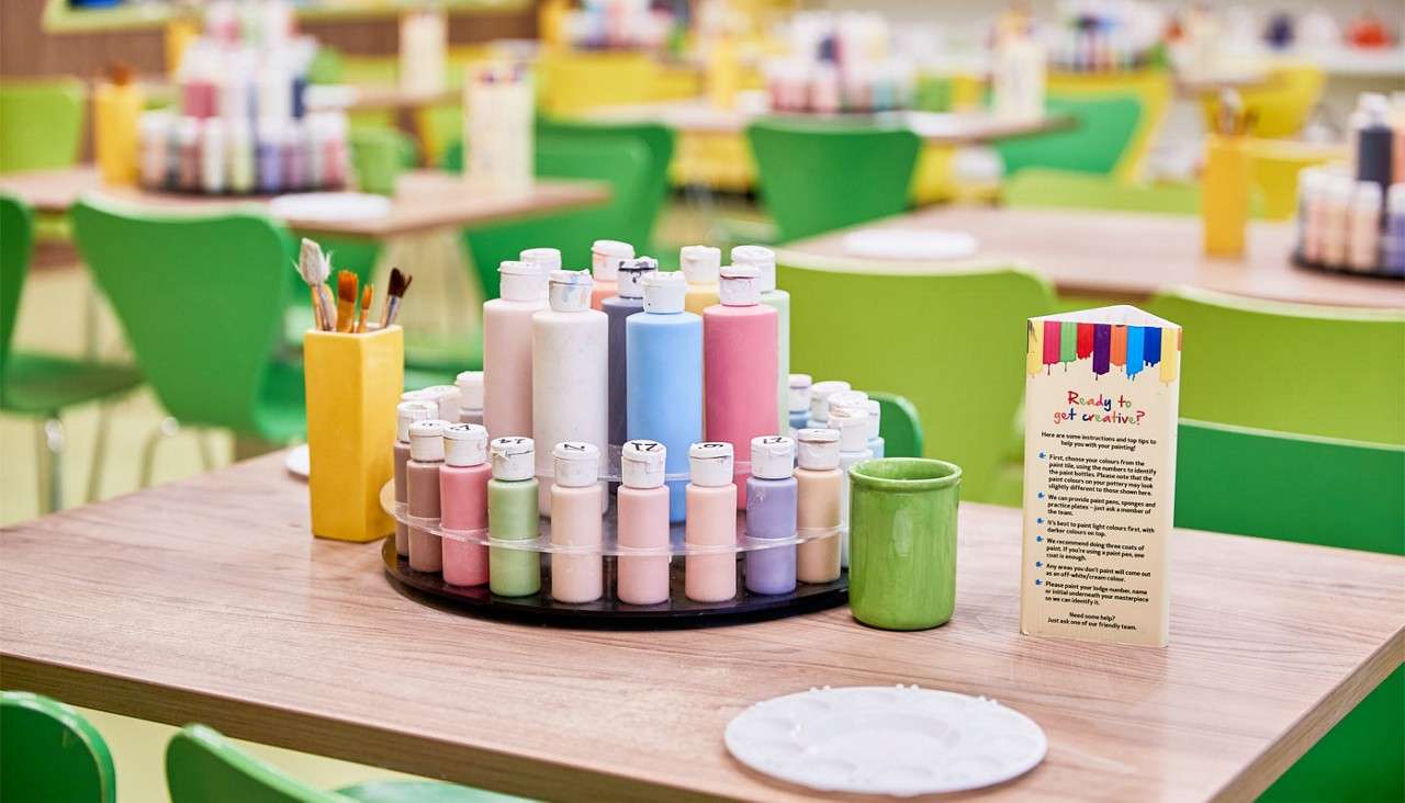 Paint bottles sit arranged on a rotating tray, accompanied by brushes, a green mug, and a paint palette, on a craft-table in a studio with green chairs. Visible text: “Ready to get creative?” Other text and bottle-cap numbers are too small to read.
