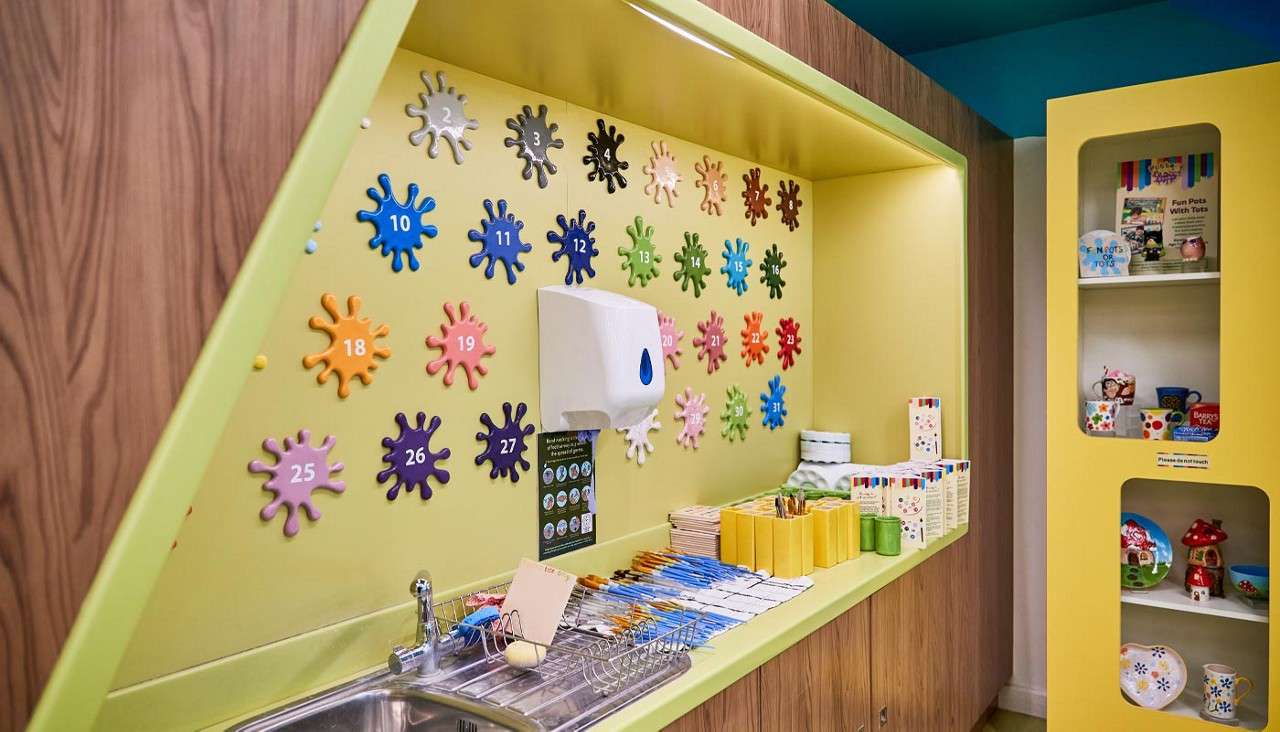 Art workstation arranges paintbrushes and tools by a sink, while a towel dispenser hangs above; colorful numbered splat tiles cover the wall; nearby shelves display painted ceramics in a bright studio.
Text: 1 2 3 4 5 6 7 8 9 10 11 12 13 14 15 16 17 18 19 20 21 22 23 24 25 26 27 28 29 30 31; Please do not touch.