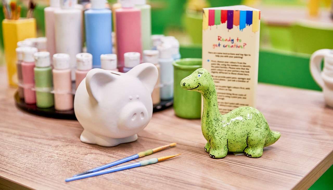 Ceramic piggy bank and green dinosaur figurine sit on a tabletop, awaiting painting with two brushes, surrounded by paint bottles and an instruction sign. Text: Ready to get creative?