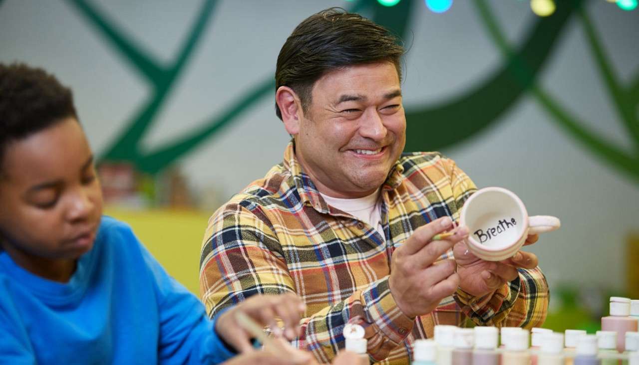 Man holds a ceramic mug, smiling while painting; inside reads “Breath”. In a craft studio, bottles of paint and a child beside him work at a table.