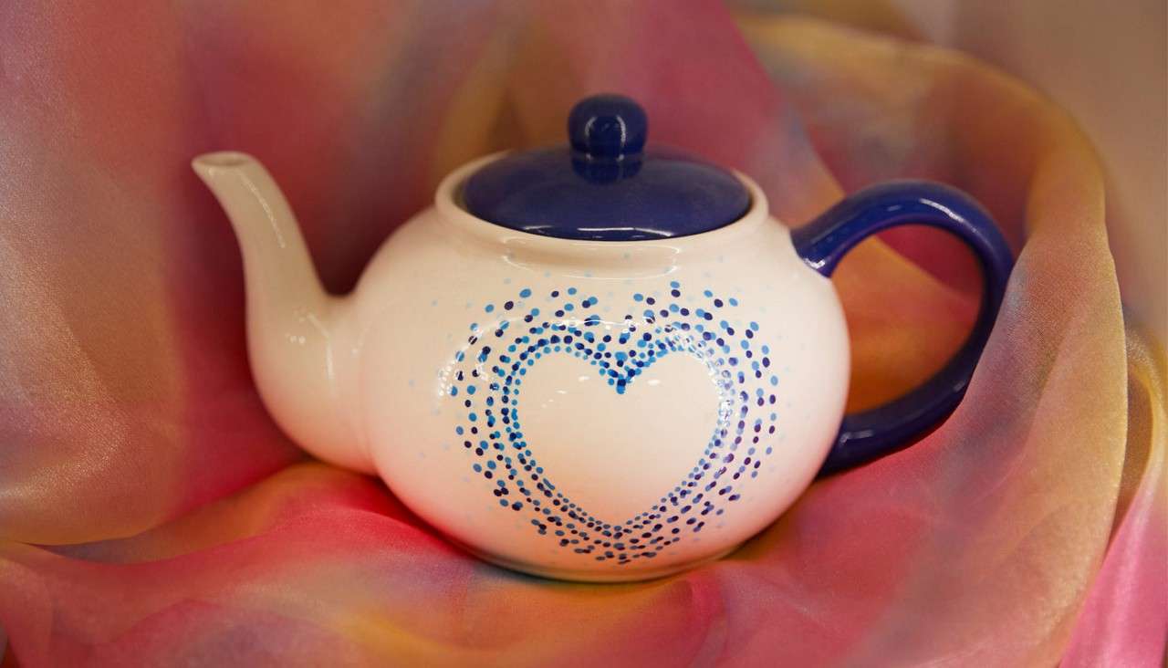 White teapot sits still, bearing a blue dotted heart motif, with navy lid and handle, nestled in draped multicolored chiffon fabric backdrop of pink, yellow, and orange.