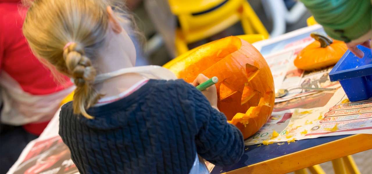 Child carves a jack-o'-lantern using a green tool at a newspaper-covered table; pumpkin pieces scattered, a small pumpkin and utensils nearby, with yellow chairs and other participants blurred in the background.