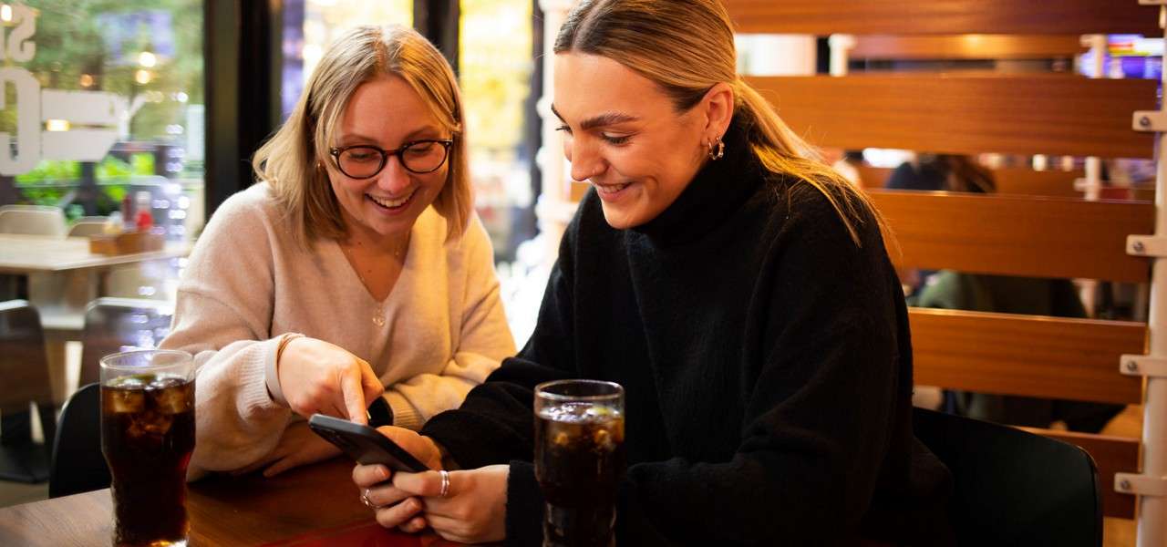 Two people smile while viewing a smartphone, one pointing at the screen, seated at a cafe table with two glasses of soda, wooden partition and windows showing greenery outside.
