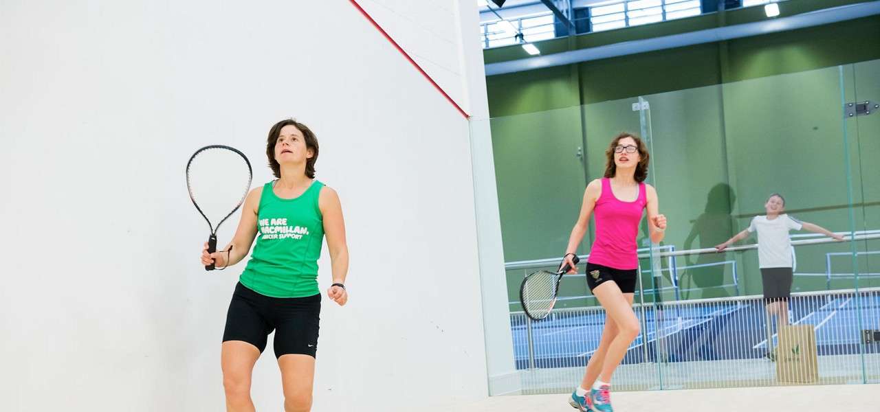 Two women with squash racquets play; one wears a green top printed “WE ARE MACMILLAN CANCER SUPPORT”. Context: indoor squash court with glass wall and a bystander outside.