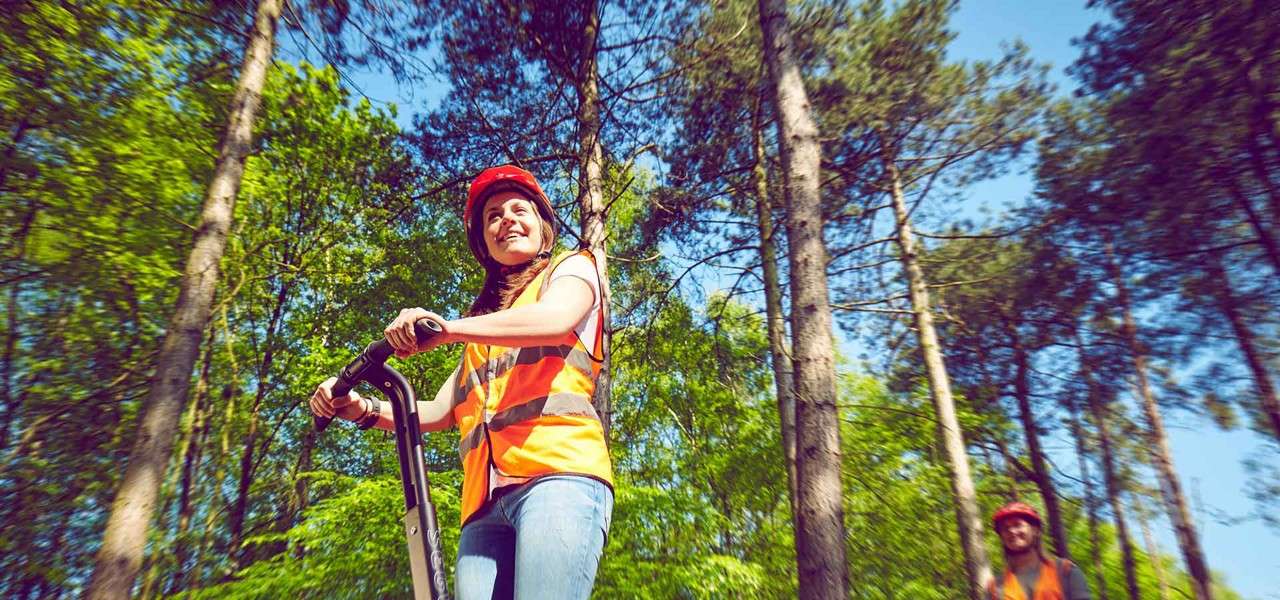 A person on a Segway rides forward, hands on handlebars, wearing a red helmet and orange safety vest, in a sunlit forest with tall trees and another rider trailing behind.