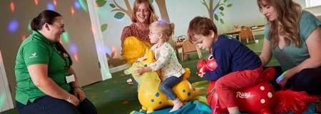 Children ride inflatable animal bouncers, bouncing and laughing, while three adults supervise in a playroom with wall tree murals and colorful light spots. Visible text: “Rody” on the red bouncer.