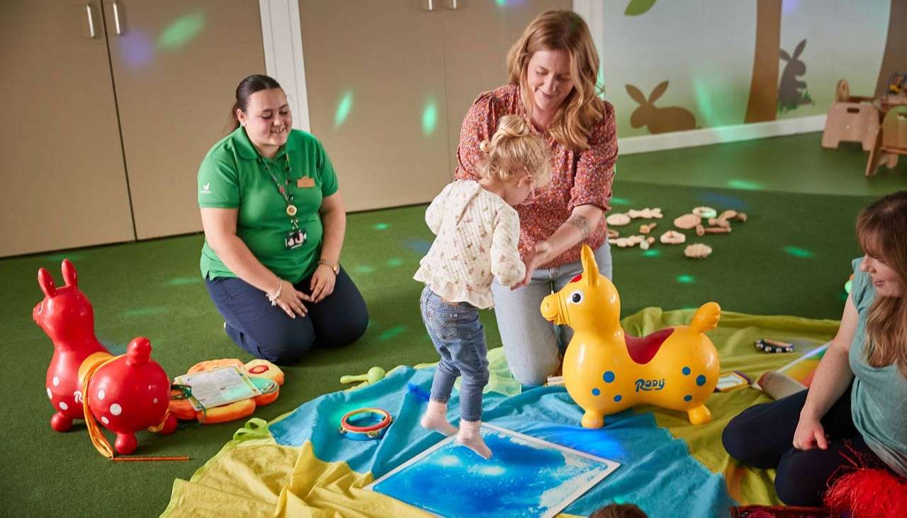 Toddler steps barefoot on a blue sensory mat while an adult steadies them; two caregivers watch. Toys and blankets surround them in a daycare room. Text: Rody.
