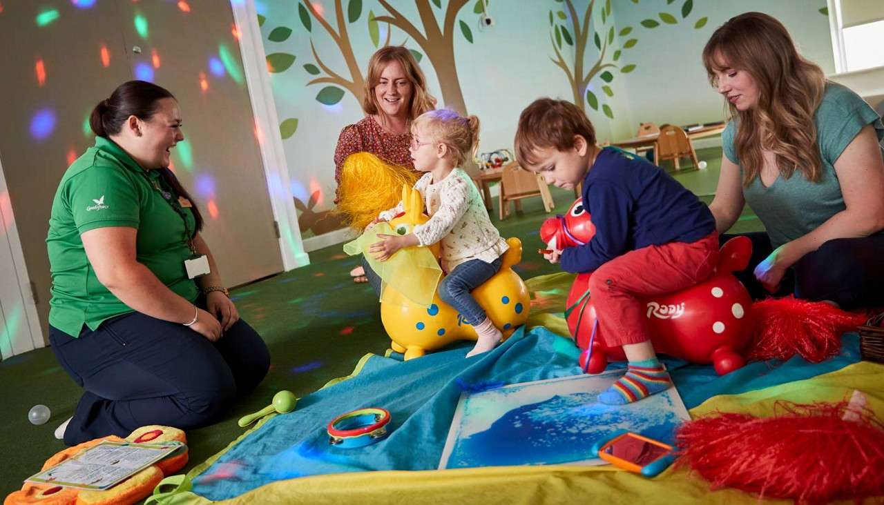 Children ride inflatable animal hoppers while adults supervise, playing with sensory toys on mats in a mural-decorated indoor playroom with colored lights. Visible text: Rody.