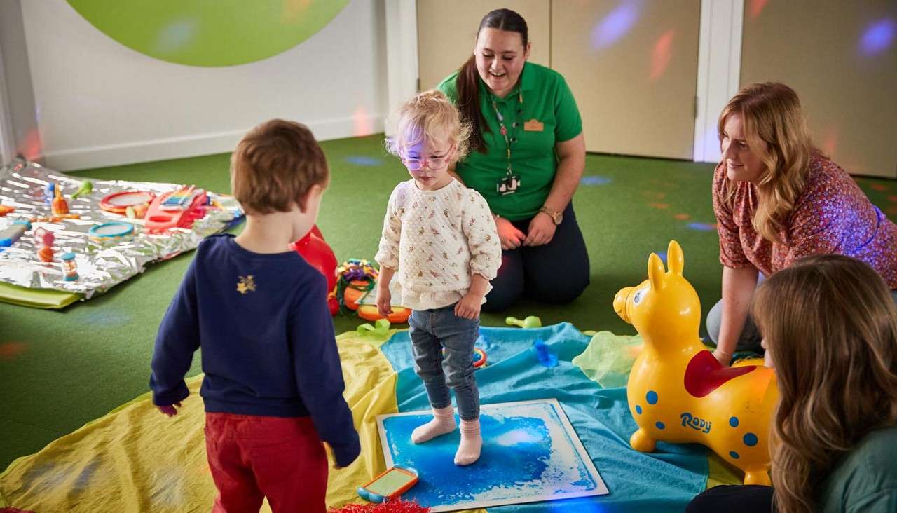 Toddler stands barefoot on a blue sensory mat, engaging another child, while three adults supervise in a green-carpeted playroom with scattered toys, blankets, and a yellow inflatable horse labeled "Rody".