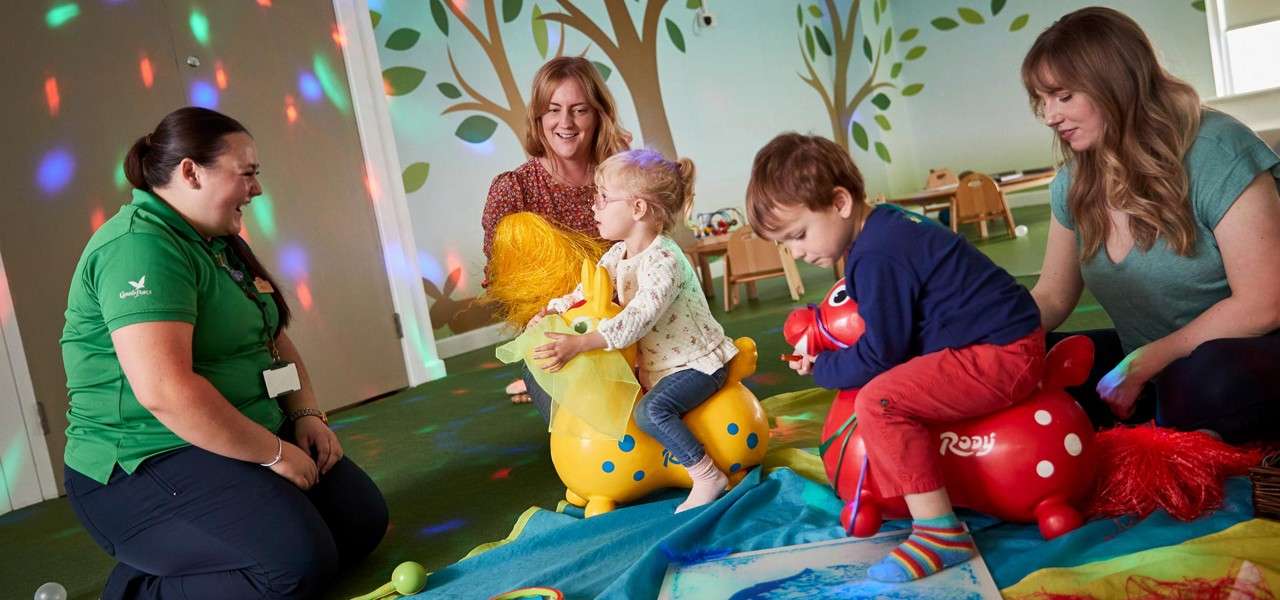 Children ride inflatable animal hoppers; adults kneel nearby, guiding and smiling; in a daycare playroom with tree murals, scattered colored light spots, mats, toys, and small tables. Text: Rody.