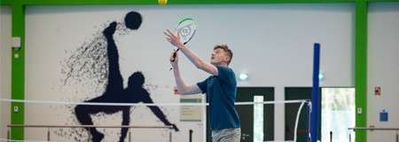 Badminton player serves a shuttlecock, reaching up with racket; inside a bright indoor court with a net, mural silhouettes of athletes on the wall, doors, and green structural beams.