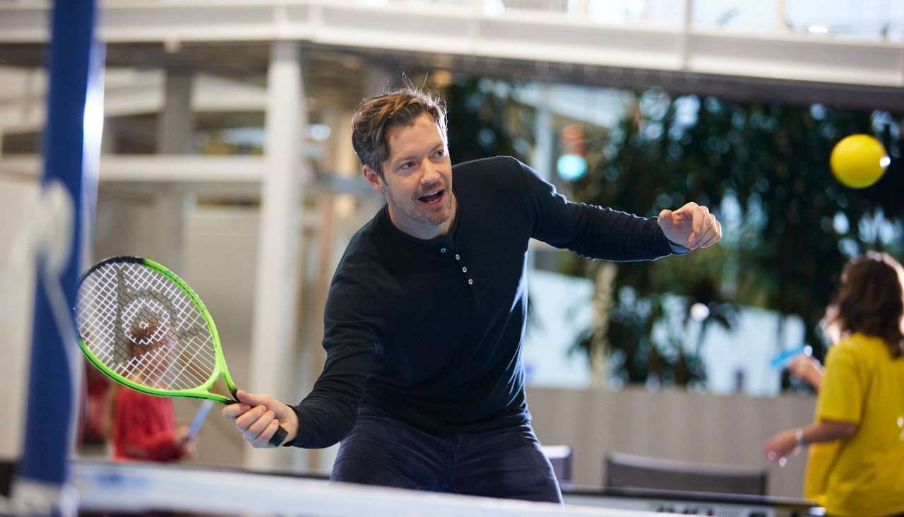 Man with a green-edged tennis racket swings to hit a yellow ball over a ping-pong table net inside a bright indoor atrium, with other people casually playing in the background.