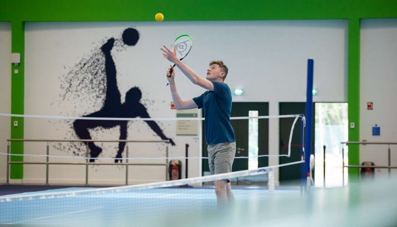 Player tosses a yellow tennis ball and raises a racket to serve on an indoor court, behind a net, with green-trimmed walls, doors, and a sports silhouette mural.