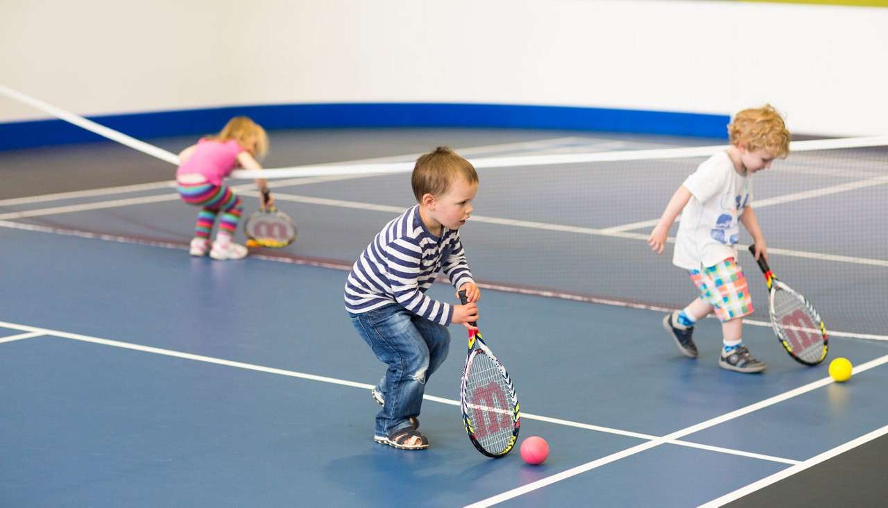 Children practice tennis, swinging small rackets to push foam balls. They crouch and step on an indoor court, with low nets and marked lines, wearing casual, colorful clothes.