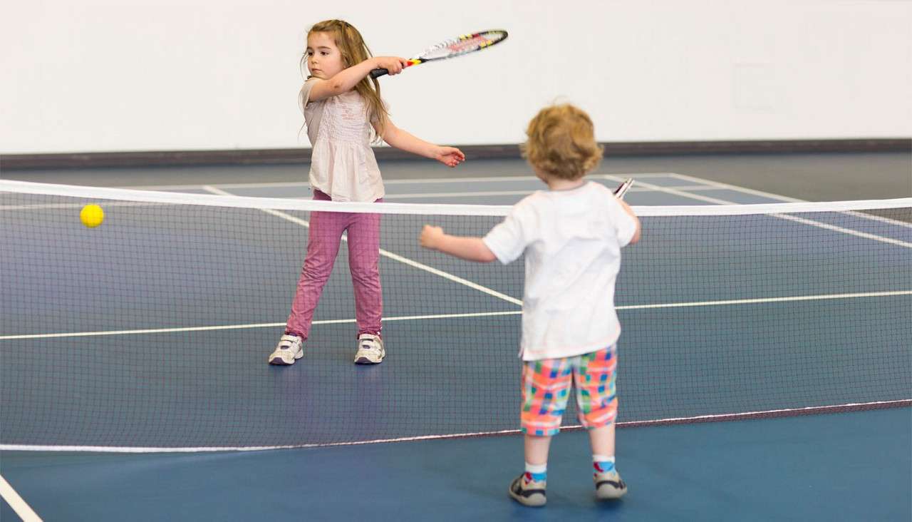 Two young children play tennis; a girl swings a racket toward a yellow ball while a toddler faces her across a low net on an indoor court.