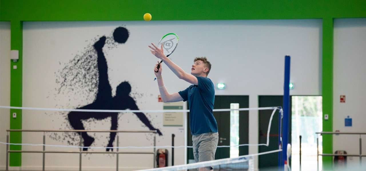 Teen player tosses a yellow ball and readies a tennis serve, standing on an indoor court beside a low net, with a large athlete mural painted on the back wall.