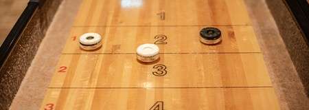 Shuffleboard pucks rest on a wooden scoring board; one white and one black sit near “2,” another white near “3.” Numbers printed: “1,” “2,” “3,” plus small red “2” markings along the sides.