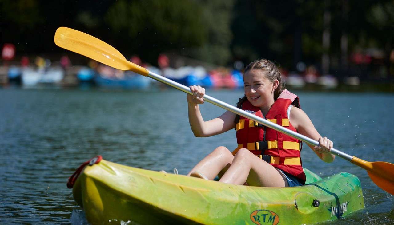Kayaker paddles a green sit-on-top kayak across a calm lake, wearing a red life jacket; background shows blurred boats near shore. Text visible on kayak: “RTM” and “MAMBO.”