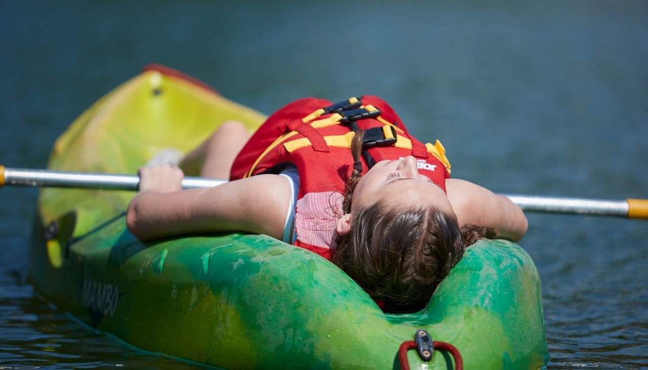 Person reclines on a green kayak, gripping a paddle across their chest, floating on calm water; wearing a red life vest. Visible text: JOBE on vest and MAMBO on kayak.