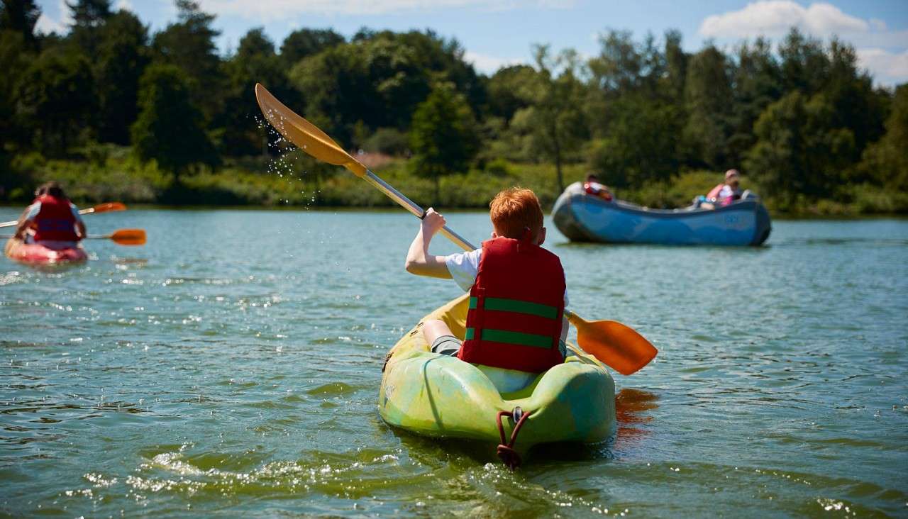 Person wearing a red life jacket paddles a green kayak on a lake; other kayaks and a raft nearby; tree-lined shore under blue sky.