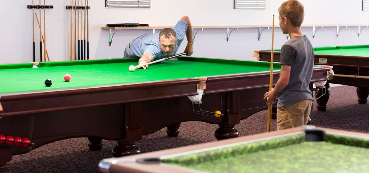 Man takes a snooker shot, leaning over a table and aiming a white ball with a rest; a boy holding a cue watches in a billiards room with multiple tables.