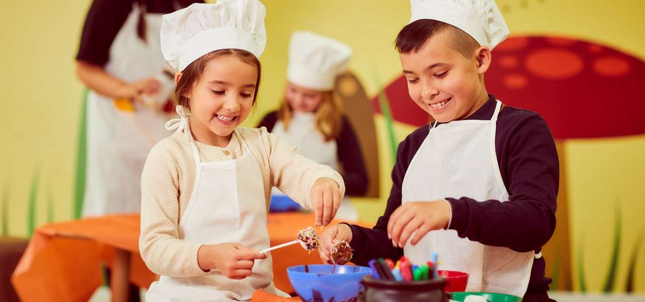 Two children wearing chef hats and aprons decorate cake pops, dipping them into bowls on a table. In the background, other kids work in a colorful classroom with mushroom-themed wall art.