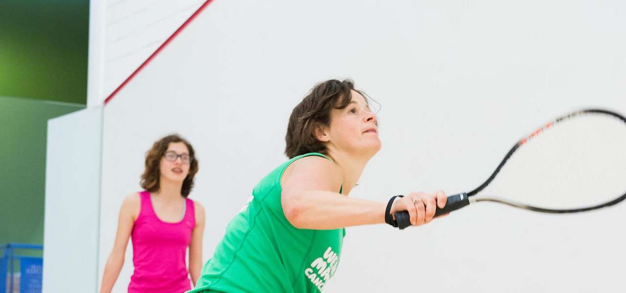 Squash player lunges forward, swinging a racket; another player watches behind, both in athletic gear on a bright, white-walled indoor court marked with a red boundary line.