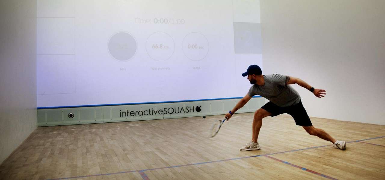Squash player lunges and swings a racket in an indoor court with a projected display. Text: Time: 0:00/1:00; 66.8 cm; 0.00 sec; Hits; Speed; 2; interactiveSQUASH.