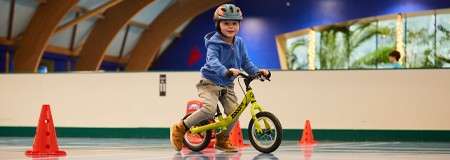 Child on a small balance bike rides through spaced orange cones, wearing a helmet, inside a bright indoor rink or gym with smooth floor and curved wooden beams.