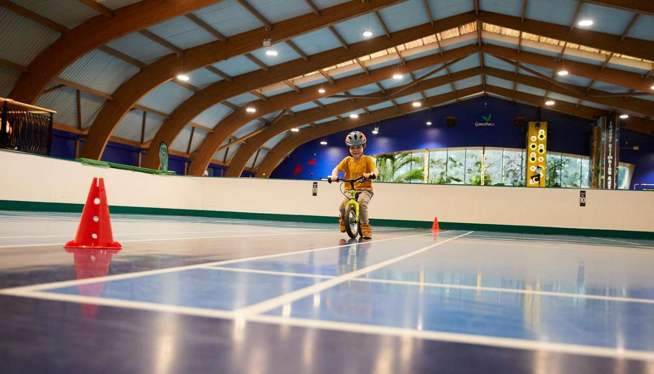 Child on a balance bike rides between orange cones on a glossy indoor court, under arched wooden beams; distant wall shows windows and climbing structures. Text: 10.