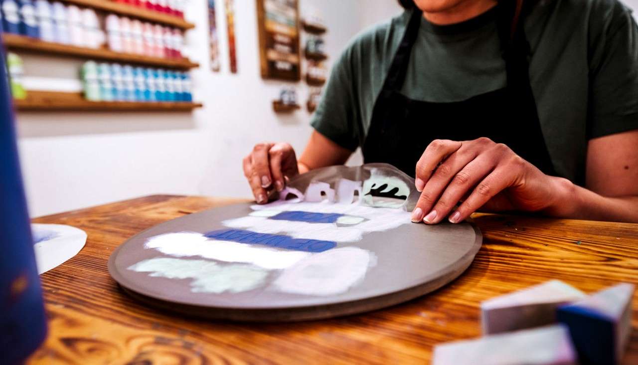 Hands shape and paint a round ceramic plate, pressing a stencil to apply abstract colored patches; context: wooden worktable in an art studio, shelves lined with organized paint bottles behind.