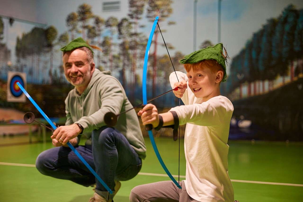 A boy and dad wearing a Robin Hood hat, dad smiles as son takes aim.