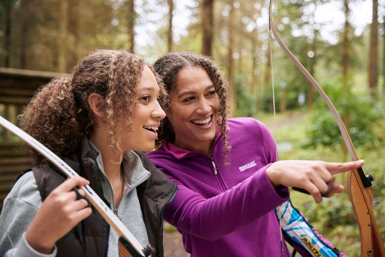 Two people laughing and pointing in the forest holding arrows. 