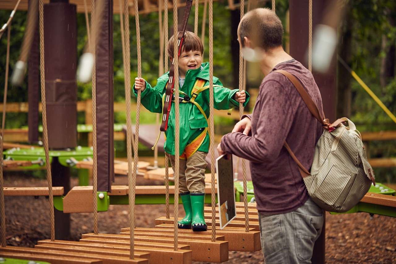 Young boy walking across a hanging bridge, with the help of an adult.