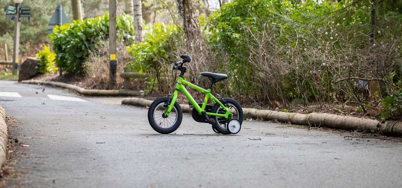 A small green children's bicycle with training wheels stands idle on a paved road, surrounded by curbed edges, trees, and bushes in a quiet park-like setting with faint crosswalk markings.